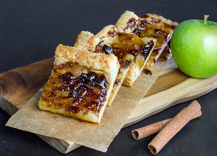 Sliced apple tart on a board alongside a fresh apple and cinnamon sticks.