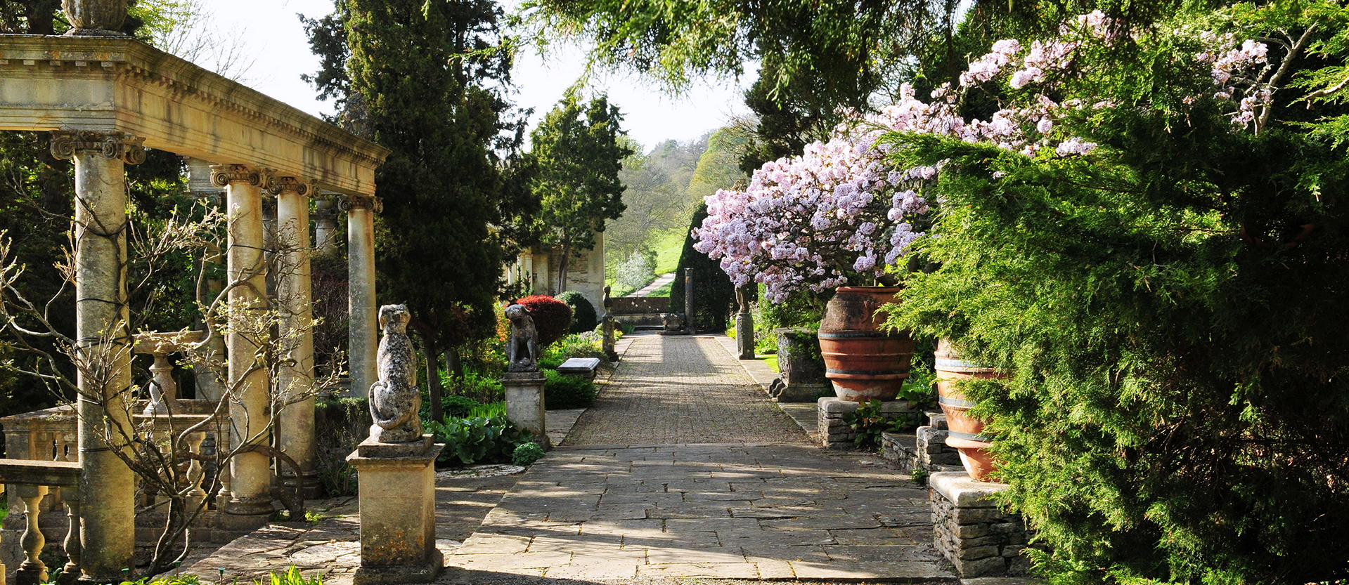 Elegant outdoor garden with stonework, a variety of florals and greenery, statues, and a marble pergola